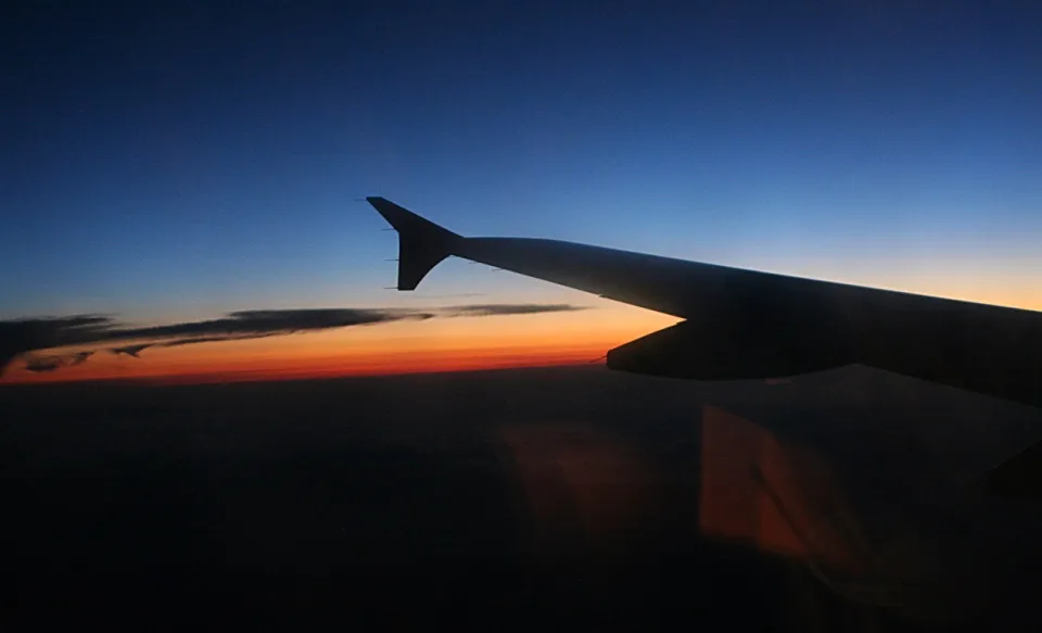 Silhouette einer Flugzeugtragfläche vor einem Himmel, der oben dunkelblau und zum Horizont hin orange leuchtet, darunter eine dunkle Wolkendecke.