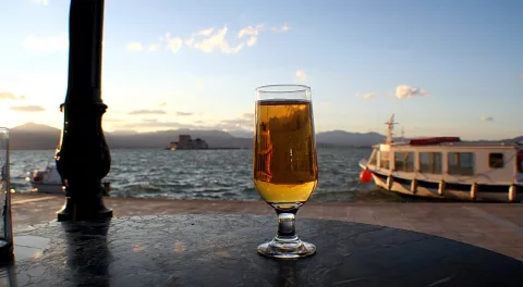 Ein Glas helles Bier steht auf einem dunklen Marmortisch am Wasser, im Hintergrund ein Ausflugsboot, eine Festung auf dem Meer und Berge unter einem Abendhimmel.