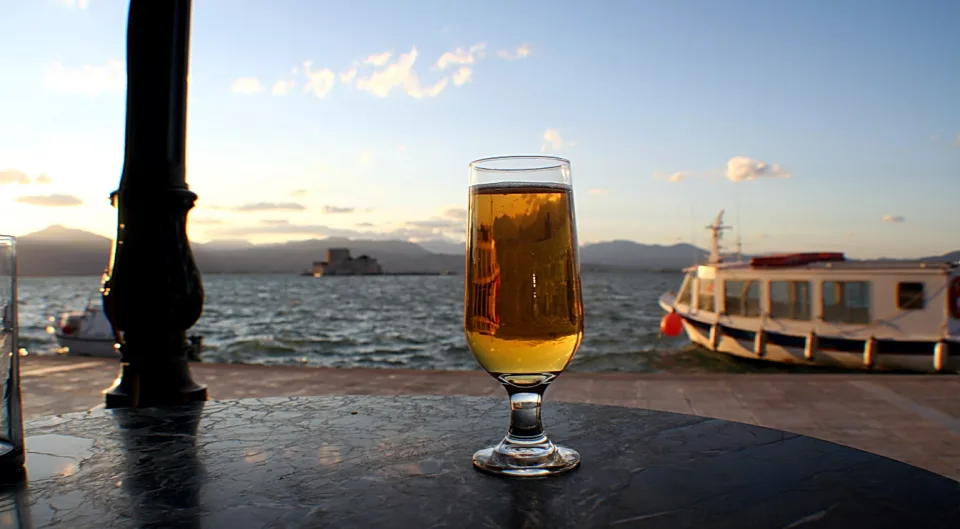 Ein Glas helles Bier steht auf einem dunklen Marmortisch am Wasser, im Hintergrund ein Ausflugsboot, eine Festung auf dem Meer und Berge unter einem Abendhimmel.