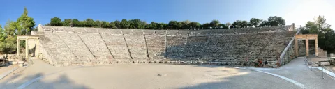 Panorama eines antiken Steintheaters mit ansteigenden Sitzreihen, einer leeren Spielfläche im Vordergrund und zwei Säulenportalen an den Seiten, davor einige sitzende Besucher.
