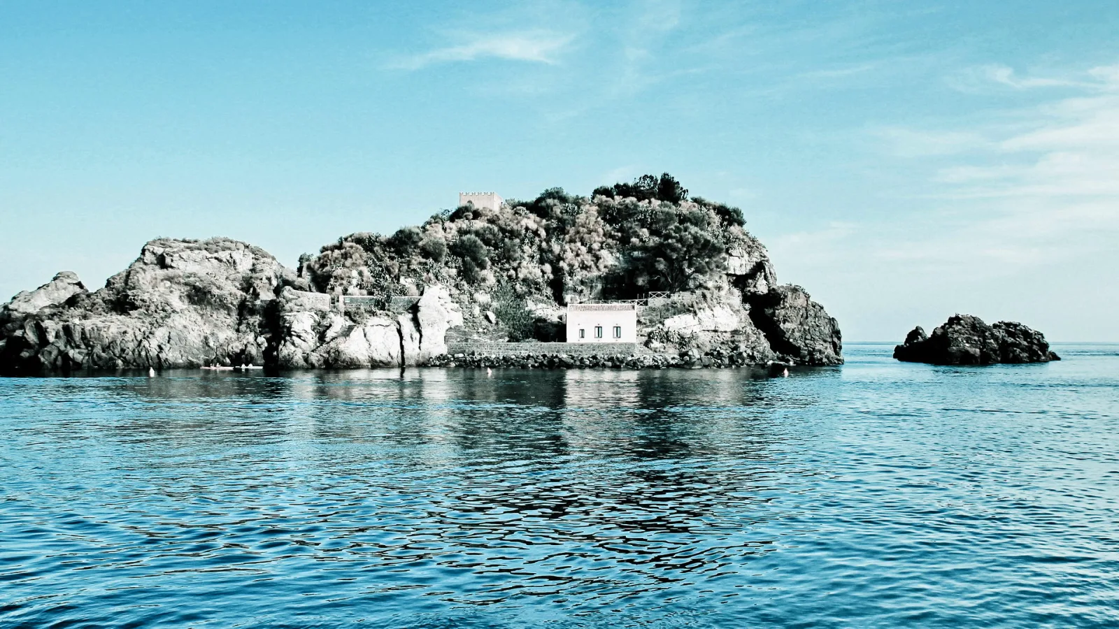 Felsige Insel mit Macchia und einem kleinen weißen Haus, davor ruhiges blaues Meer unter klarem Himmel