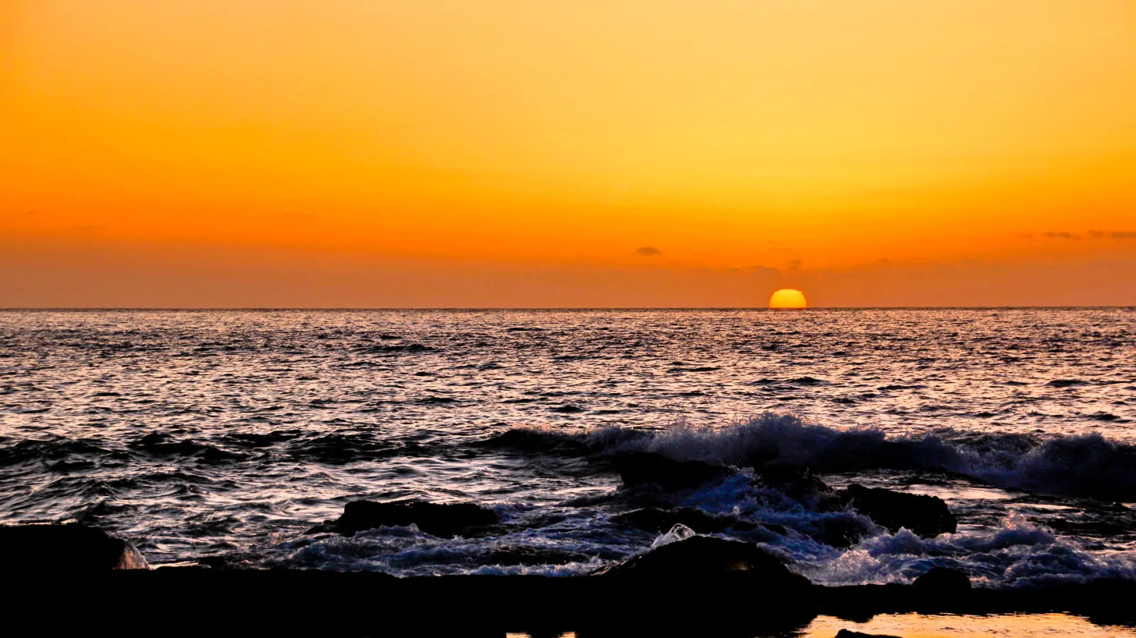 Die Sonne sinkt knapp über dem Horizont, der Himmel leuchtet orange, im Vordergrund brechen Wellen an dunklen Felsen.