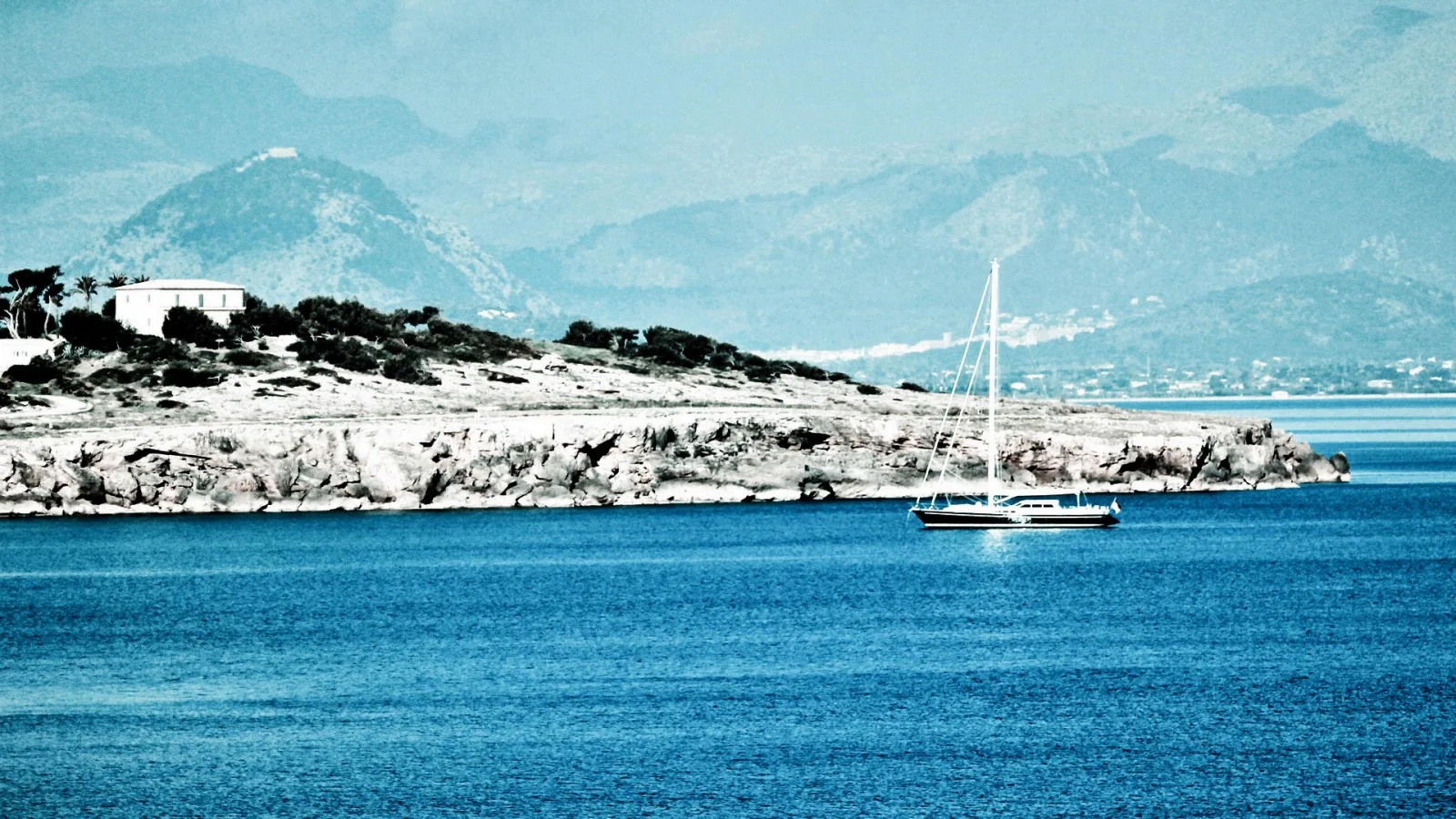 Ein weißes Segelboot liegt auf tiefblauem Meer vor einer hellen, felsigen Küste mit einem Gebäude und Bäumen, dahinter blaue Berge im Dunst.