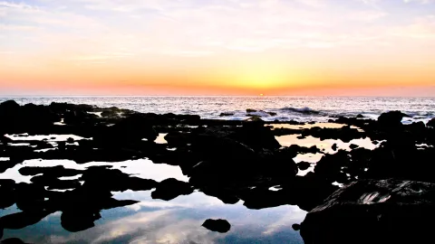 Dunkle Lavafelsen mit Gezeitentümpeln im Vordergrund, dahinter das Meer mit Wellen und die untergehende Sonne über dem Horizont vor orangefarbenem Himmel.