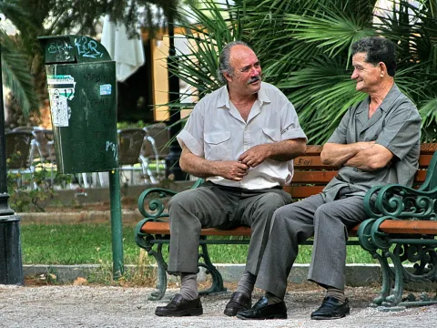 Zwei ältere Männer sitzen auf einer grünen Parkbank und unterhalten sich, rechts davor steht ein grüner Mülleimer, im Hintergrund sind Palmen zu sehen.