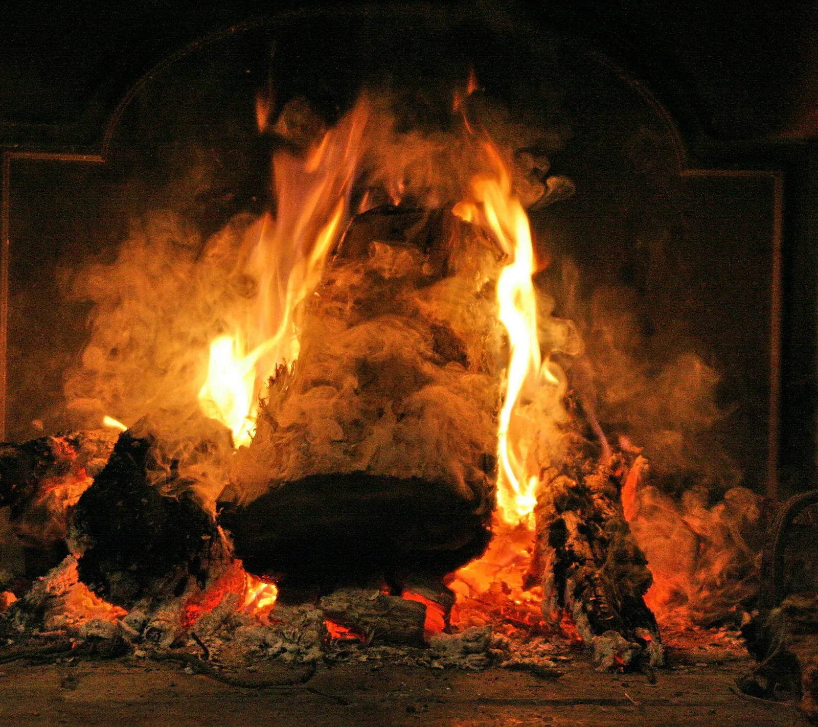 Brennender Holzscheit im offenen Kamin, Flammen schlagen seitlich hoch, dazwischen dichter Qualm und glühende Holzkohle am Boden.