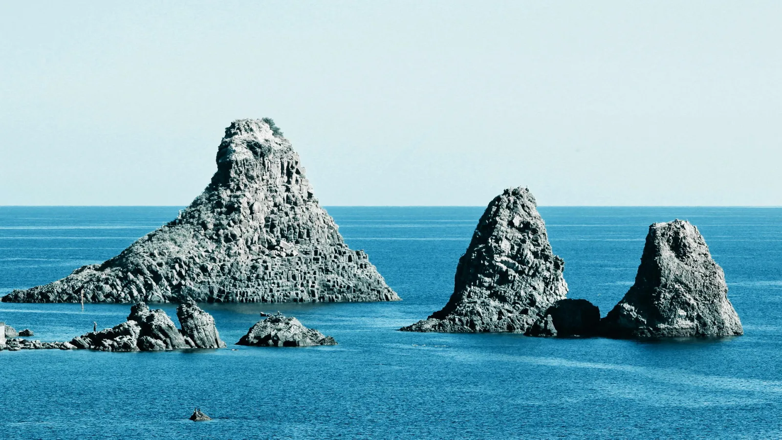 Mehrere dunkle Felsnadeln und ein breiter Basaltfelsen ragen aus blauem, ruhigem Meer, darüber ein klarer, hellblauer Himmel.