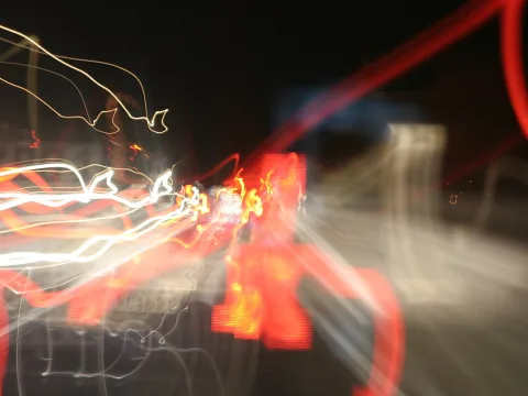 Verwackelte Langzeitaufnahme nachts: weiße, orangefarbene und rote Lichtspuren ziehen sich diagonal über ein dunkles Bild, rechts verschwimmt ein rot leuchtendes Gebäude.