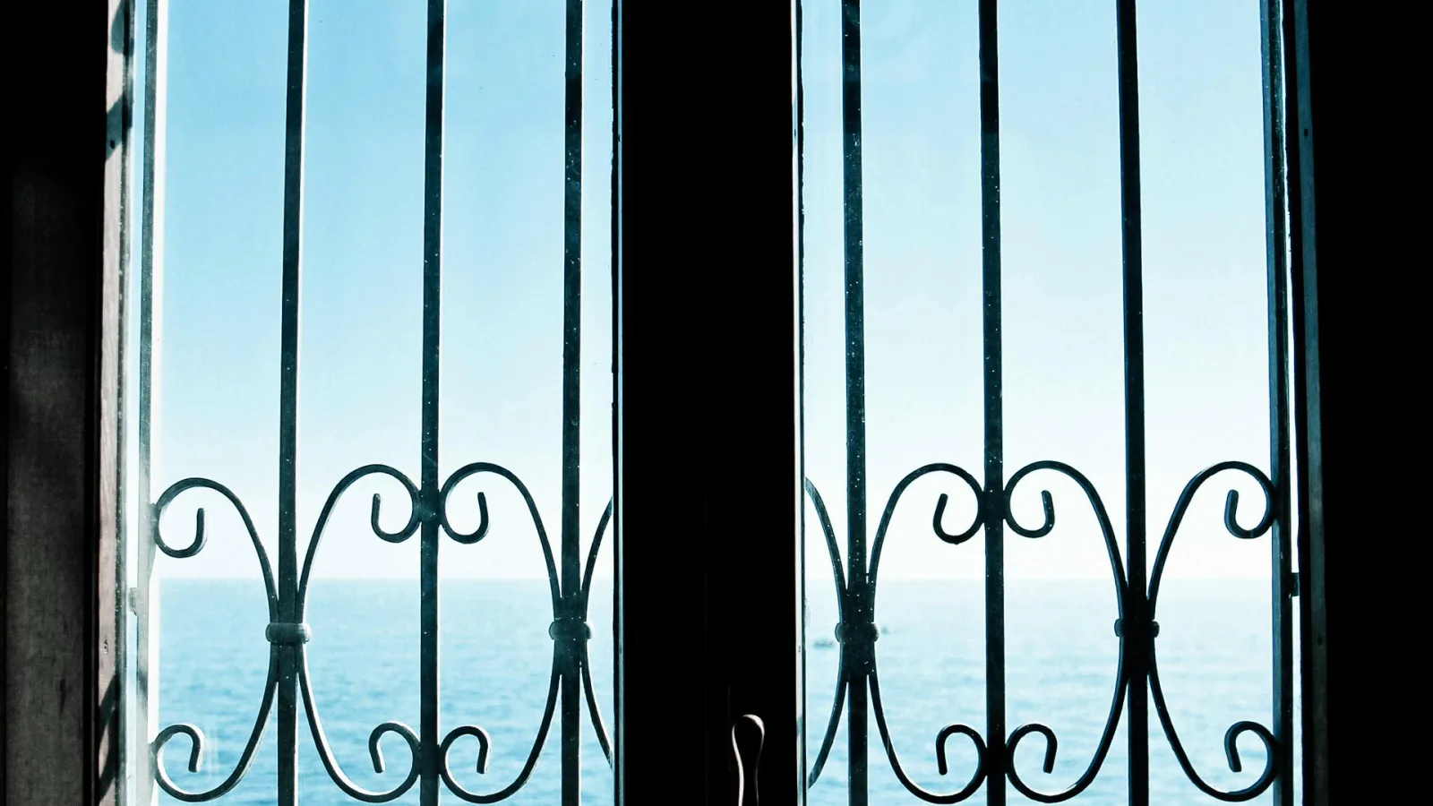 Dunkles Fenster mit vertikalen Eisenstäben und geschwungenen Schmiedeornamenten, dahinter blauer Himmel und das Meer am Horizont