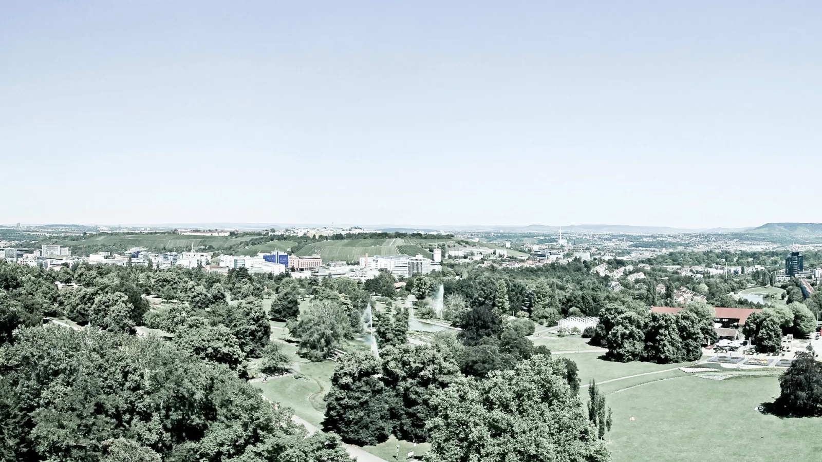 Weitblick über einen Stadtpark mit dichten Baumkronen, zwei Fontänen und einer grünen Wiese im Vordergrund, dahinter Stadtbebauung und Hügel unter klarem Himmel.