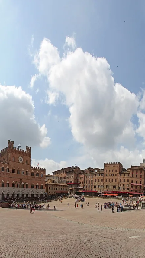 Großer gepflasterter Platz in Siena mit Palazzo Pubblico und Turm links, Menschen und Cafés mit roten Sonnenschirmen, blauer Himmel mit Wolken.