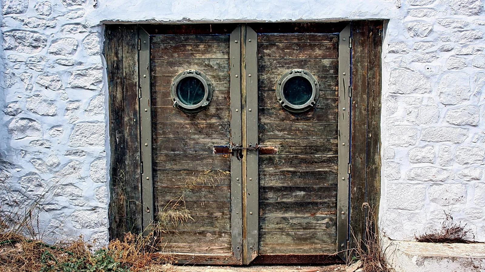 Zwei alte Holztüren mit runden Bullaugen und Metallbeschlägen, mit Kette verschlossen, in einer weiß getünchten Steinmauer.