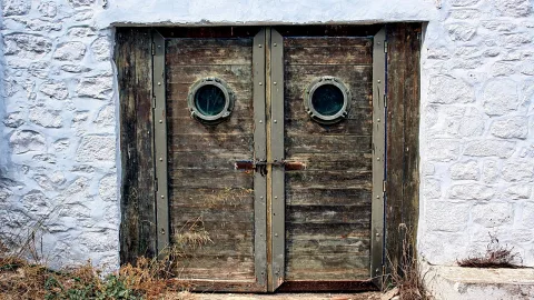 Zwei alte Holztüren mit runden Bullaugen und Metallbeschlägen, mit Kette verschlossen, in einer weiß getünchten Steinmauer.