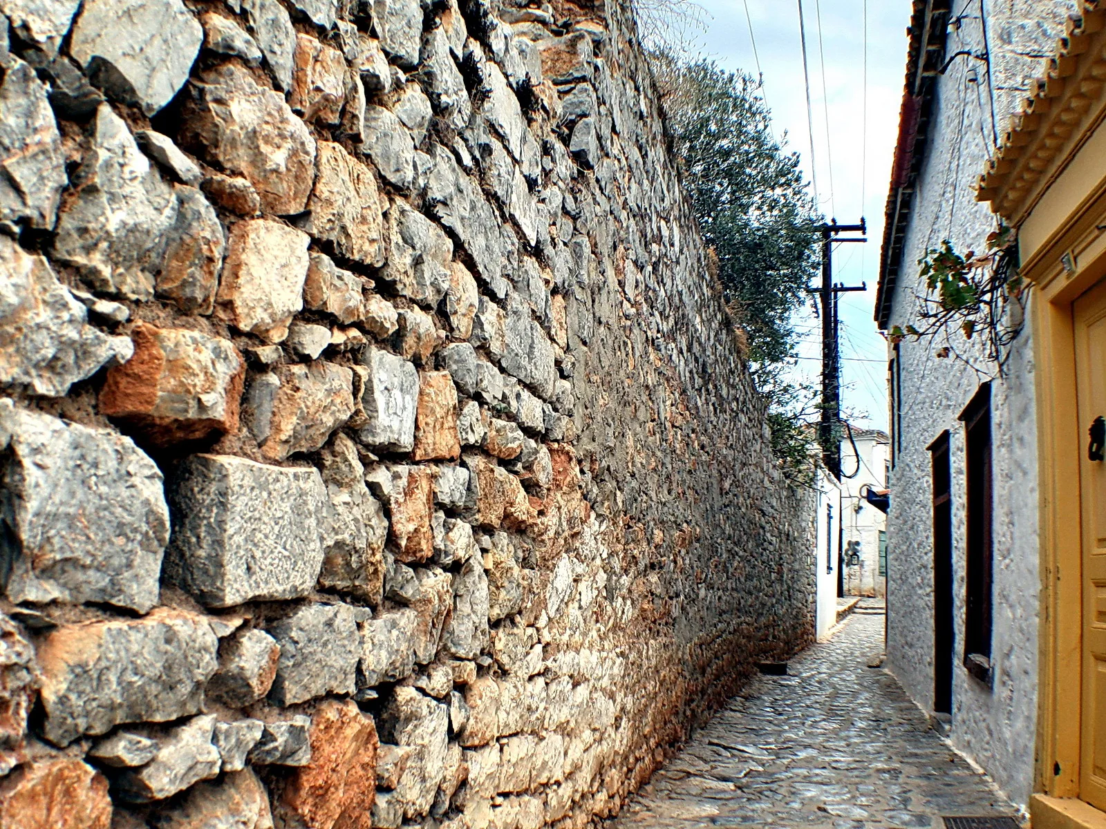 Enge Kopfsteinpflastergasse zwischen einer hohen Bruchsteinmauer links und einer weiß verputzten Hauswand mit gelber Tür rechts, darüber Stromleitungen und ein Olivenbaum.