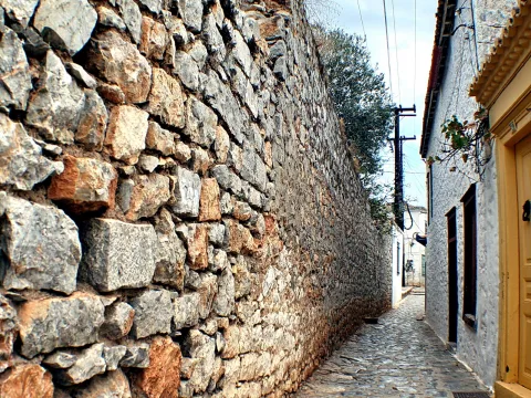 Enge Kopfsteinpflastergasse zwischen einer hohen Bruchsteinmauer links und einer weiß verputzten Hauswand mit gelber Tür rechts, darüber Stromleitungen und ein Olivenbaum.