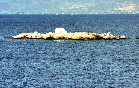Felseninsel mit einem weißen Häuschen und einer Antenne im blauen Meer, dahinter bewaldete Hügel und kleine Segelboote.