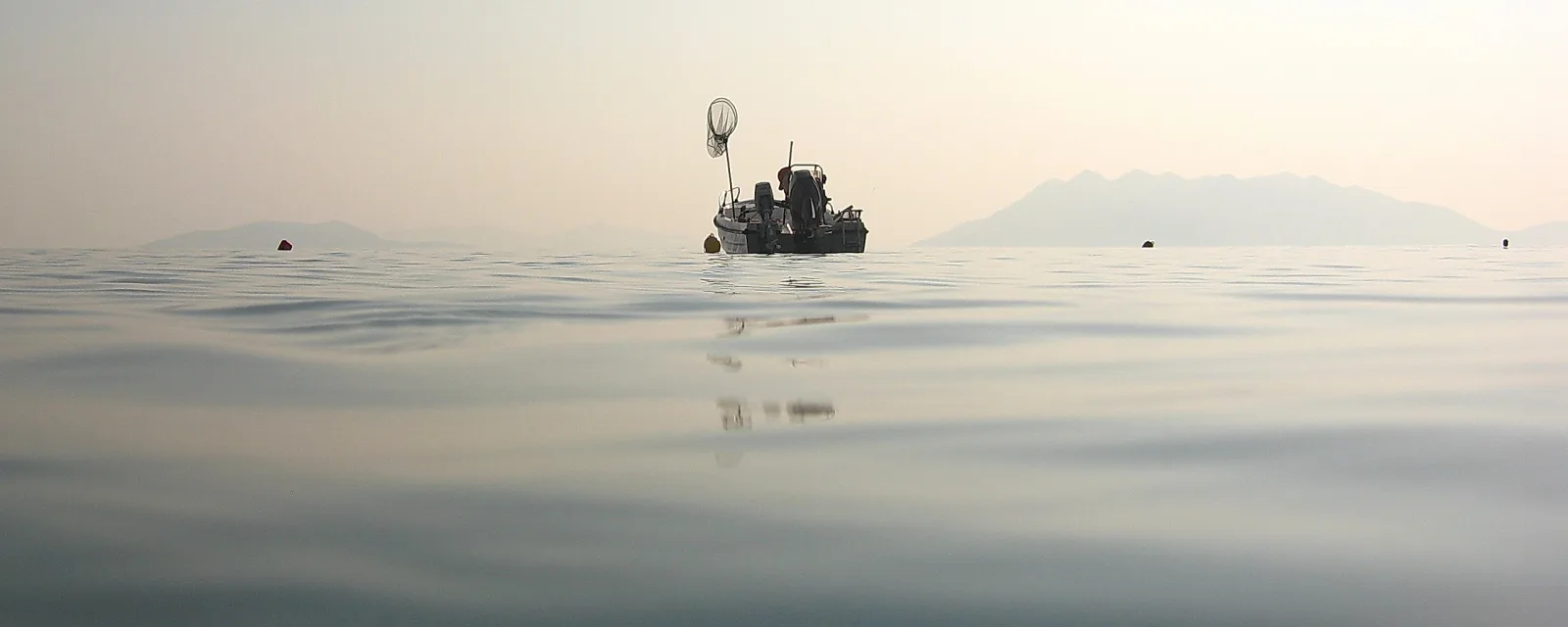Ein kleines Fischerboot mit Netz auf glatter Wasseroberfläche, im Dunst sind Berge am Horizont zu sehen.