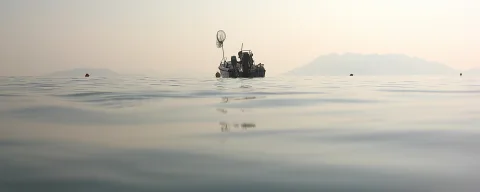 Ein kleines Fischerboot mit Netz auf glatter Wasseroberfläche, im Dunst sind Berge am Horizont zu sehen.