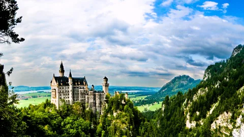 Schloss Neuschwanstein mit hellen Türmen auf einem bewaldeten Felsen, im Hintergrund Berge, Seen und grüne Wiesen unter einem bewölkten Himmel.