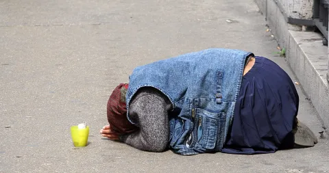 Eine Person kniet mit gesenktem Kopf auf grauem Asphalt, daneben steht ein gelber Becher.