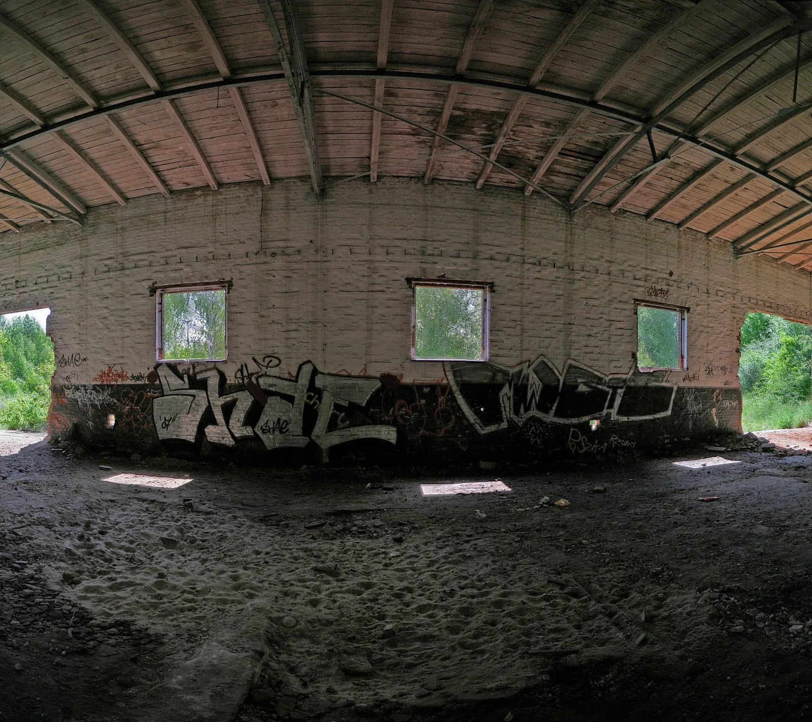 Innenraum einer verlassenen Halle mit gewölbter Holzdecke, weiß verputzter Backsteinwand mit Graffiti und drei Fenstern, durch die Bäume zu sehen sind, sandiger Boden mit Schutt.