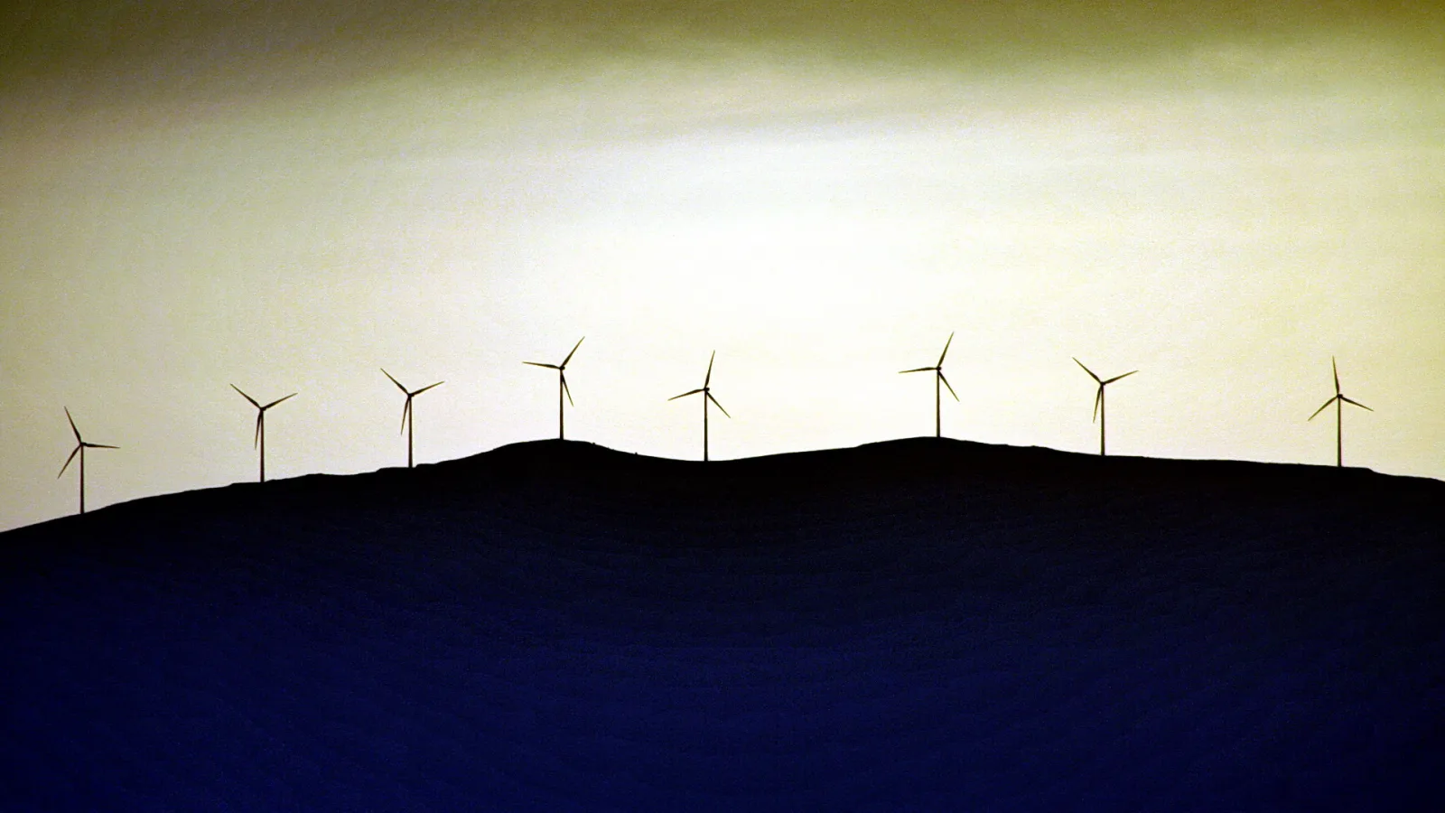 Silhouetten von acht Windkraftanlagen auf einem dunklen Hügelkamm vor einem gelb-grauen Abendhimmel.