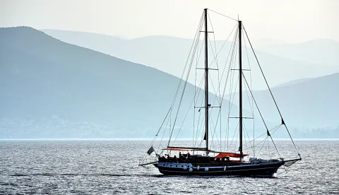 Ein zweimastiges Segelschiff mit dunklem Rumpf und roter Persenning fährt bei Dunst über ruhiges Wasser, im Hintergrund liegen blau getönte Berge.