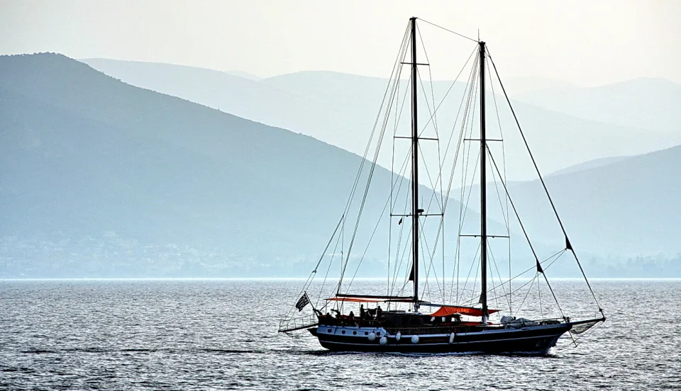 Ein zweimastiges Segelschiff mit dunklem Rumpf und roter Persenning fährt bei Dunst über ruhiges Wasser, im Hintergrund liegen blau getönte Berge.