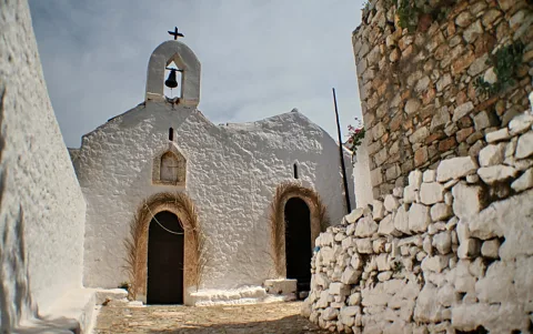 Eine weiß getünchte Kapelle mit Glockenturm, Glocke und Kreuz steht am Ende einer engen Gasse, links und rechts begrenzt von weißen und Steinmauern.