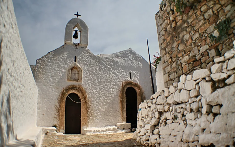 Eine weiß getünchte Kapelle mit Glockenturm, Glocke und Kreuz steht am Ende einer engen Gasse, links und rechts begrenzt von weißen und Steinmauern.