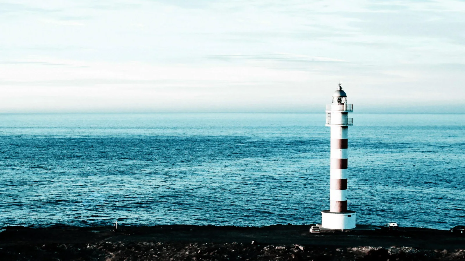 Weiß-rot gestreifter Leuchtturm auf einer dunklen Küste, dahinter ruhiges blaues Meer und heller Himmel.