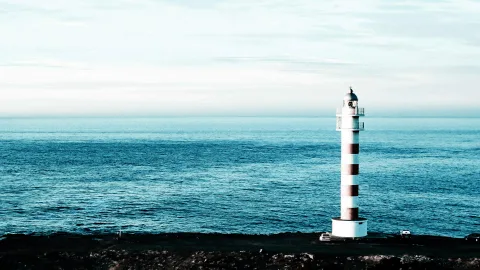 Weiß-rot gestreifter Leuchtturm auf einer dunklen Küste, dahinter ruhiges blaues Meer und heller Himmel.