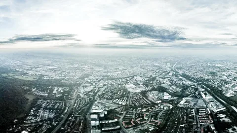 Luftaufnahme einer weitläufigen Stadt mit Straßen, Wohnvierteln und Industriegebäuden, darüber ein heller Himmel mit Wolkenbändern und dunstigem Horizont.