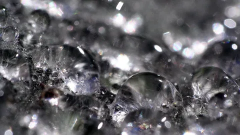 Makroaufnahme vieler kleiner Wassertropfen auf einer dunklen, glitzernden Oberfläche, mit hellen Lichtreflexen und Unschärfe im Hintergrund.