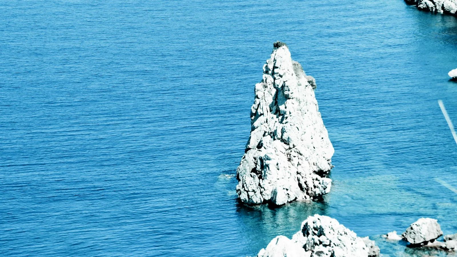 Ein einzelner grauer Felsen ragt aus blauem Meer, daneben liegen kleinere Felsen am unteren und rechten Bildrand.