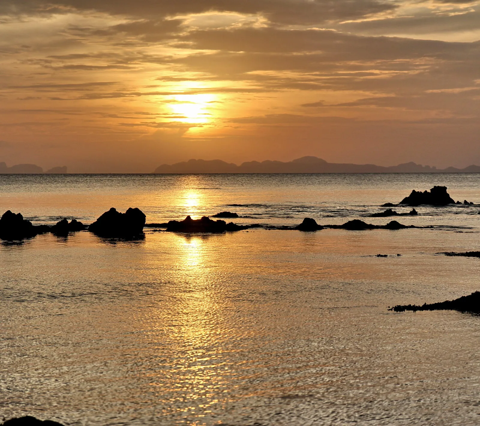 Sonnenuntergang über dem Meer, dunkle Felsen ragen aus dem flachen Wasser, im Hintergrund zeichnen sich Inseln am Horizont ab.