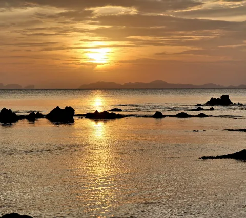 Sonnenuntergang über dem Meer, dunkle Felsen ragen aus dem flachen Wasser, im Hintergrund zeichnen sich Inseln am Horizont ab.