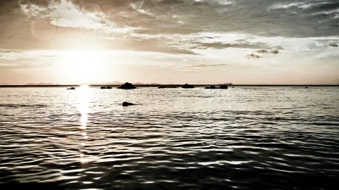 Ruhige Meeresoberfläche mit leichten Wellen im Abendlicht, die Sonne tief am Horizont, einige Felsen im Wasser und ein bewölkter Himmel.