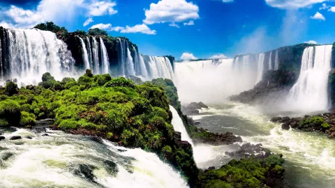 Panoramablick auf die Iguazú-Wasserfälle mit weißer Gischt, grün bewachsenen Felsinseln und blauem Himmel mit Wolken.