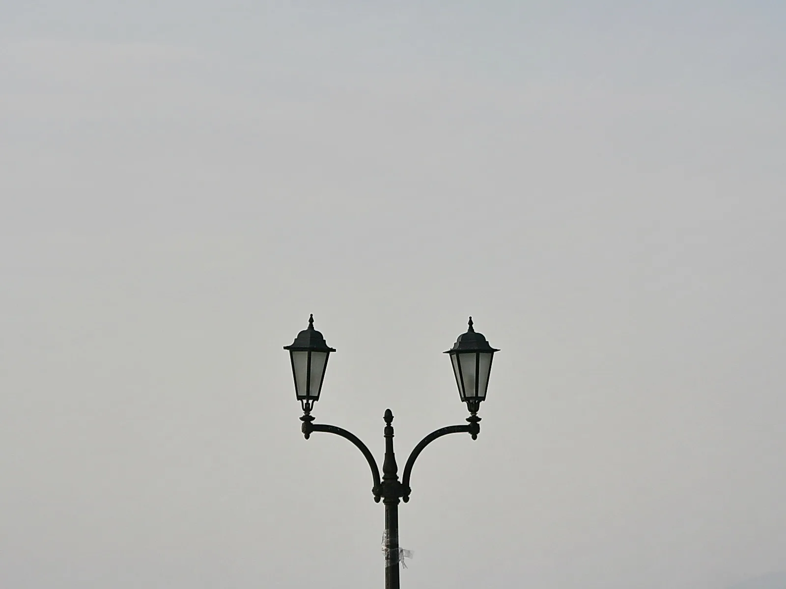 Schwarze Doppelstraßenlaterne mit zwei Laternen auf geschwungenen Armen vor einem gleichmäßig grauen Himmel.