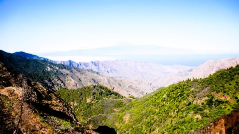 Blick über grün bewaldete Bergtäler und zerklüftete Hänge, im Hintergrund ein Berg über dem Dunst am Horizont