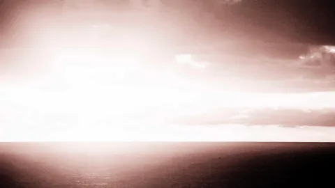Helles Sonnenlicht hinter Wolken über einer weiten, dunklen Meeresfläche, der Horizont verläuft waagerecht durch die Bildmitte, in warmen Sepiatönen.