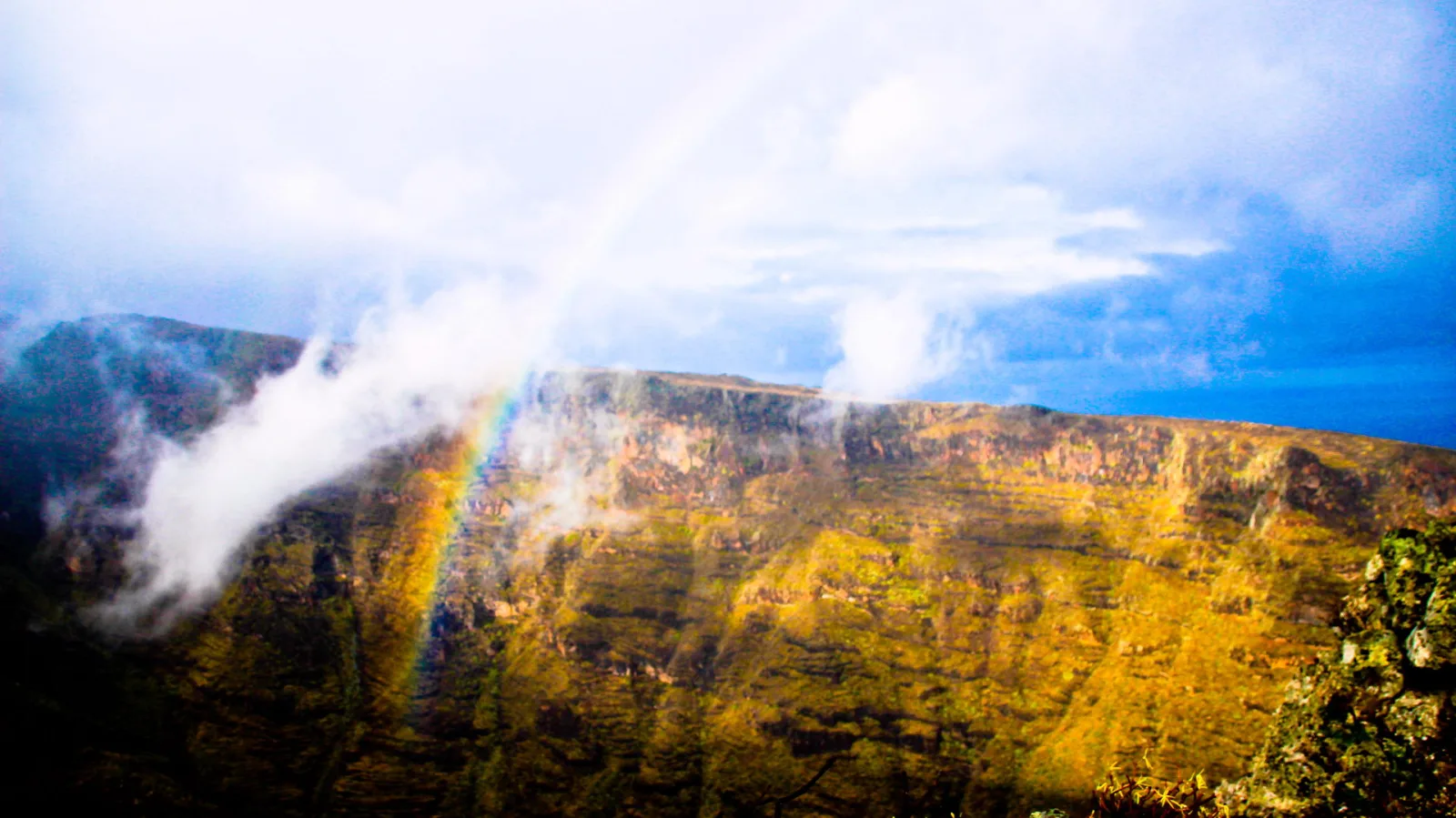 Steil abfallende, gelbgrün bewachsene Felswand mit Schichtung, davor ein Regenbogen im Nebel, darüber blauer Himmel mit weißen Wolken.