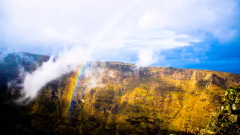 Steil abfallende, gelbgrün bewachsene Felswand mit Schichtung, davor ein Regenbogen im Nebel, darüber blauer Himmel mit weißen Wolken.