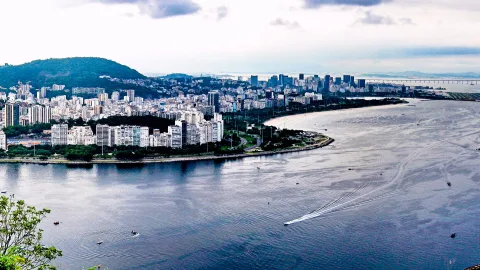 Blick von einer Anhöhe über eine Bucht mit Hochhäusern, Küstenpromenade und Brücke in Rio de Janeiro, auf dem Wasser ein Motorboot mit weißer Heckspur.
