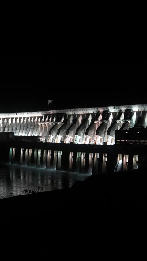 Breiter, hell beleuchteter Staudamm mit Betonpfeilern und Schleusenöffnungen vor schwarzem Nachthimmel, davor dunkles Wasser.
