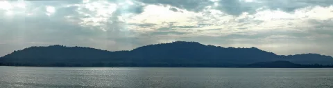 Dunkle, bewaldete Hügelkette hinter einer ruhigen Wasserfläche unter bewölktem Himmel mit durchbrechenden Sonnenstrahlen.