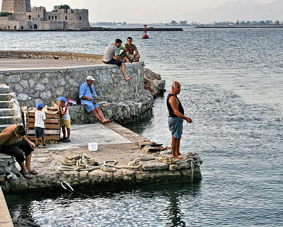 Mehrere Männer und zwei Kinder am Hafen einer Steinmole. Ein Mann im Unterhemd steht am Wasser, andere sitzen auf der Mauer. Im Hintergrund liegt eine Festung auf einer Insel, davor das ruhige Meer.