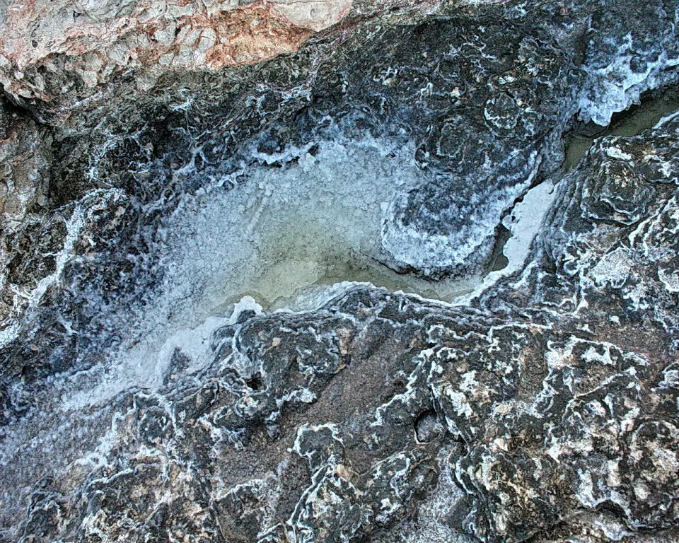 Dunkle Felsoberfläche mit weißer Salzkruste und einer flachen, ausgetrockneten Pfütze in der Mitte.