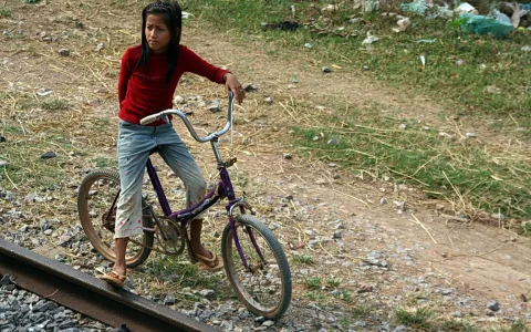Ein Mädchen in rotem Pullover und Jeans steht auf einem lila Fahrrad neben einem Eisenbahngleis, im Hintergrund Gras und Müll.